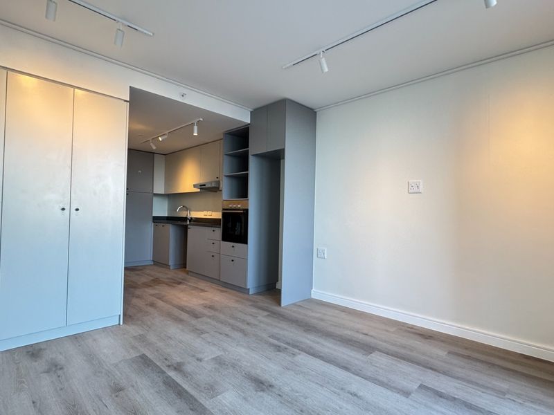Modern open-plan living area featuring light laminate flooring, track lighting, and a sleek, neutral-toned kitchen with built-in appliances and ample storage.