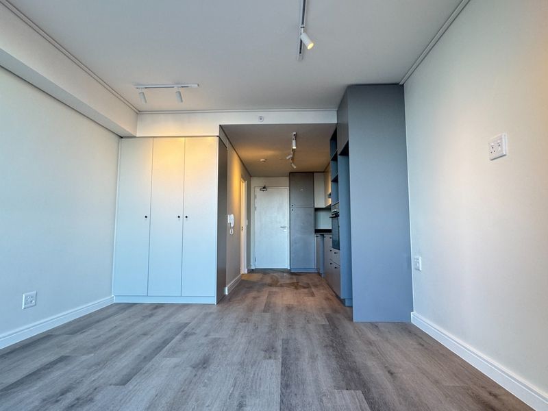 A modern, empty open-plan living area featuring sleek built-in cabinetry, laminate flooring, and contemporary track lighting in a well-maintained apartment.