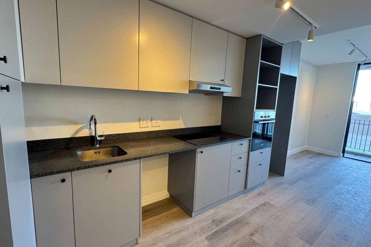 Modern kitchen featuring sleek cabinetry, stone countertops, integrated oven, electric hob, and track lighting, flowing into an open-plan living area with balcony access.