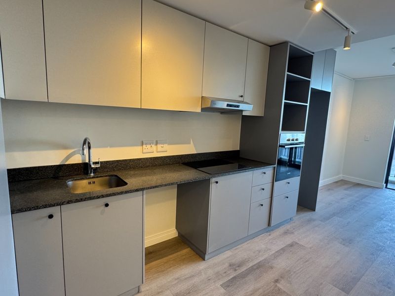 Modern kitchen featuring sleek cabinetry, stone countertops, integrated oven, electric hob, and track lighting, flowing into an open-plan living area with balcony access.