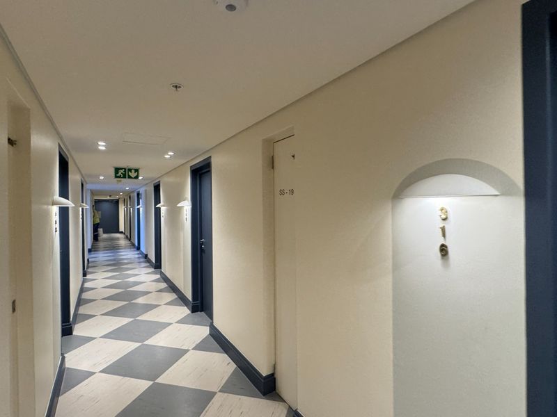 A well-maintained apartment building hallway featuring checkered floor tiles, recessed wall lighting, and clearly marked unit doors.