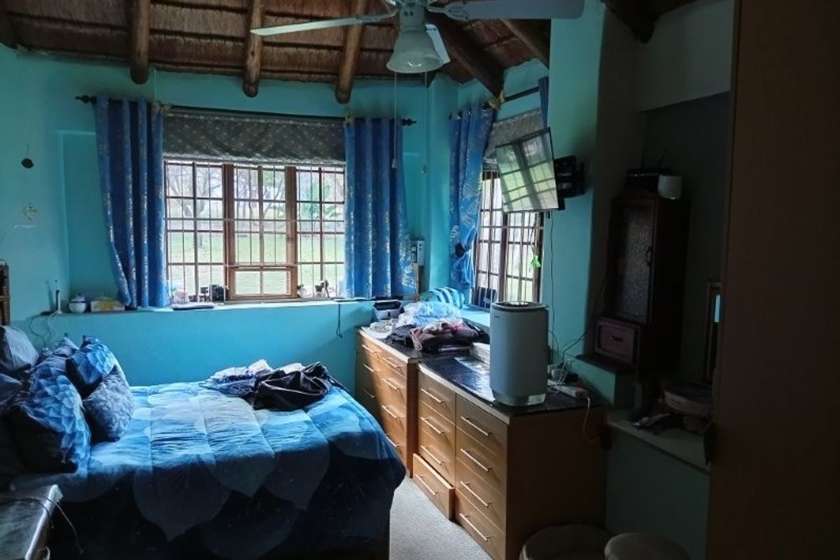 A bedroom featuring a traditional thatched roof with exposed timber rafters, a ceiling fan, and timber-framed windows overlooking the farm grounds.
