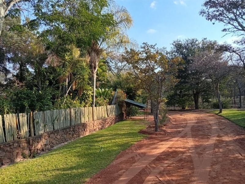 A well-maintained farm garden featuring a stone retaining wall, timber fencing, and a dirt driveway surrounded by mature trees and lush greenery.