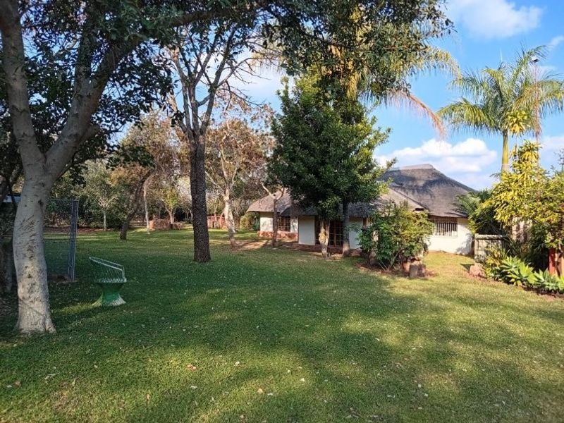 A view of the rear garden featuring a thatched roof dwelling surrounded by mature trees and a well-maintained lawn.