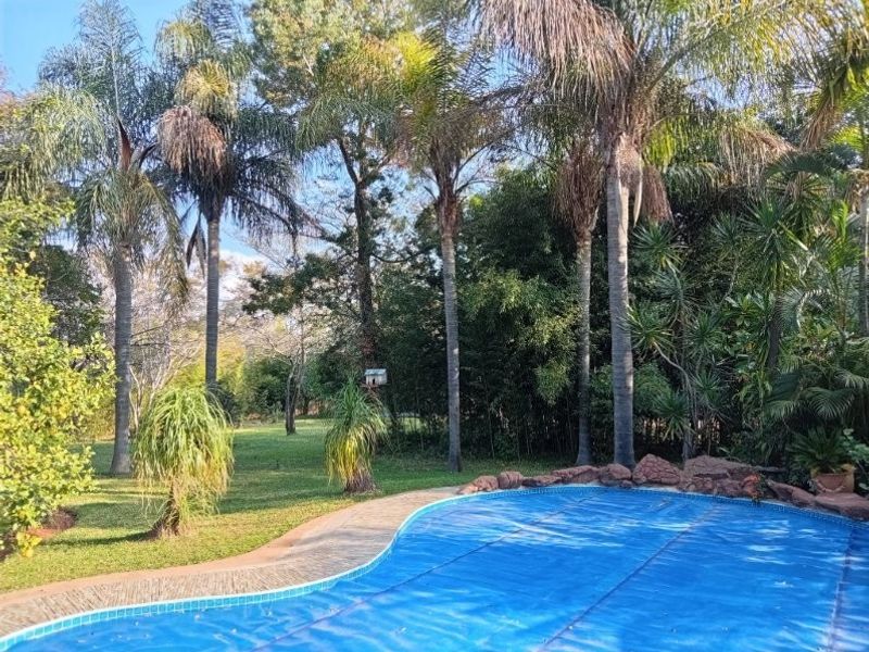 A well-maintained swimming pool area featuring a blue pool cover, paved surround, and established garden landscaping with mature palm trees.