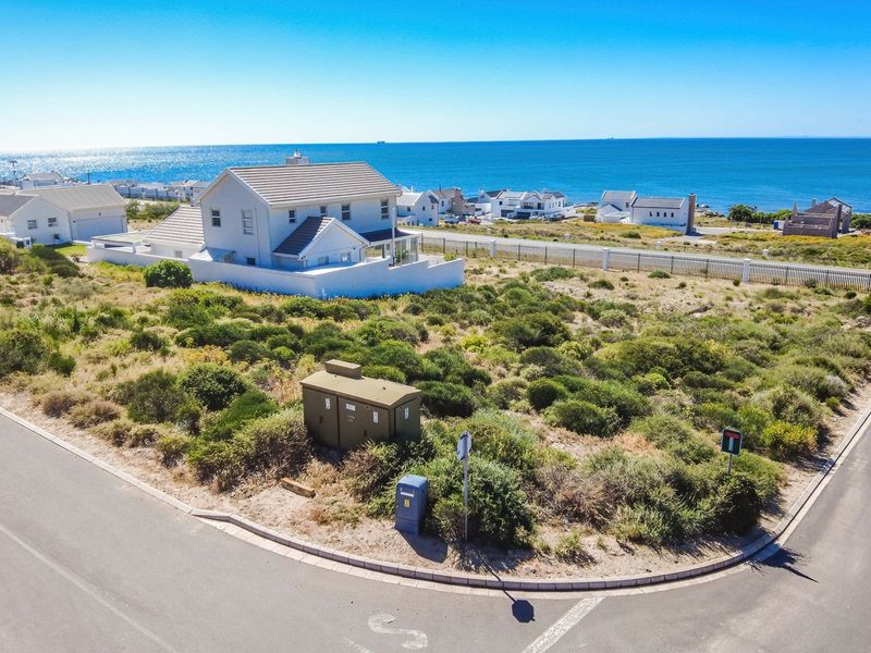 Aerial view of a vacant residential corner plot featuring native coastal vegetation, an electrical transformer box, and paved street access with ocean views.