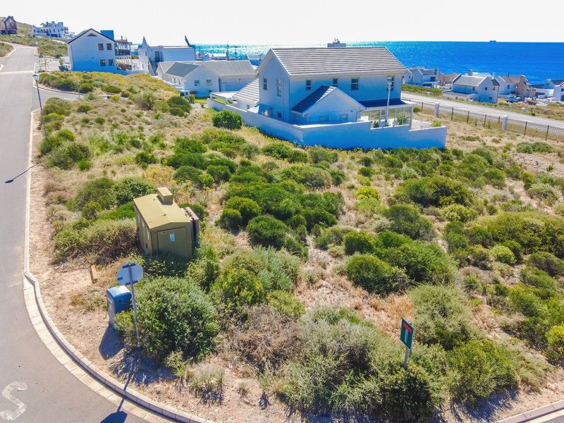An aerial view of a vacant residential plot featuring natural coastal vegetation, adjacent to a paved road and established residential housing.