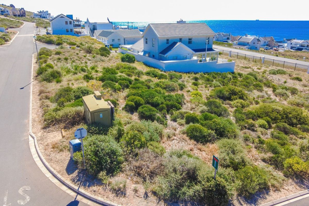 An aerial view of a vacant residential plot featuring natural coastal vegetation, adjacent to a paved road and established residential housing.