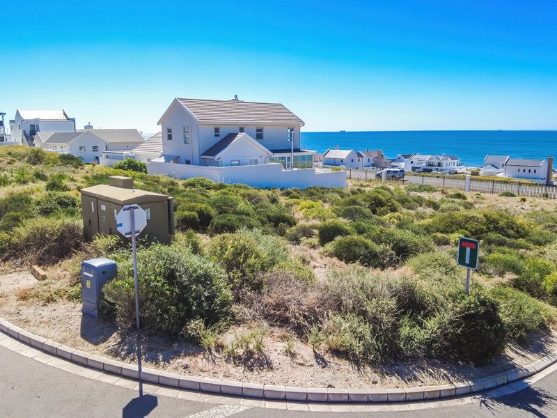 An aerial view of a vacant residential plot featuring native vegetation, a corner curb, and nearby electrical infrastructure with coastal views.