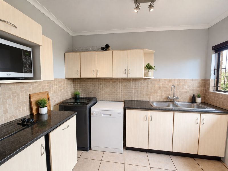 A functional scullery featuring light wood cabinetry, dark laminate countertops, a double basin sink, and tiled backsplash with space for laundry appliances.