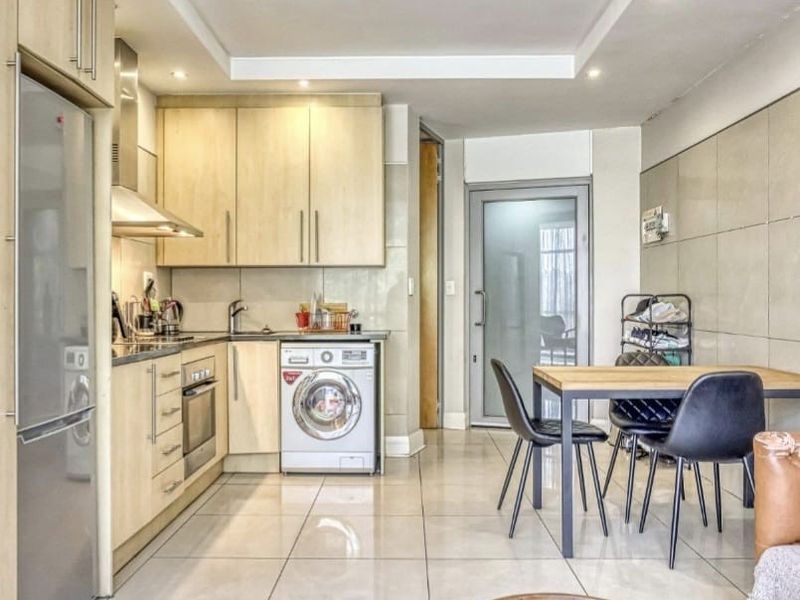 Modern open-plan kitchen and dining area featuring light wood cabinetry, integrated appliances, tiled flooring, and recessed lighting with a glass door leading outside.