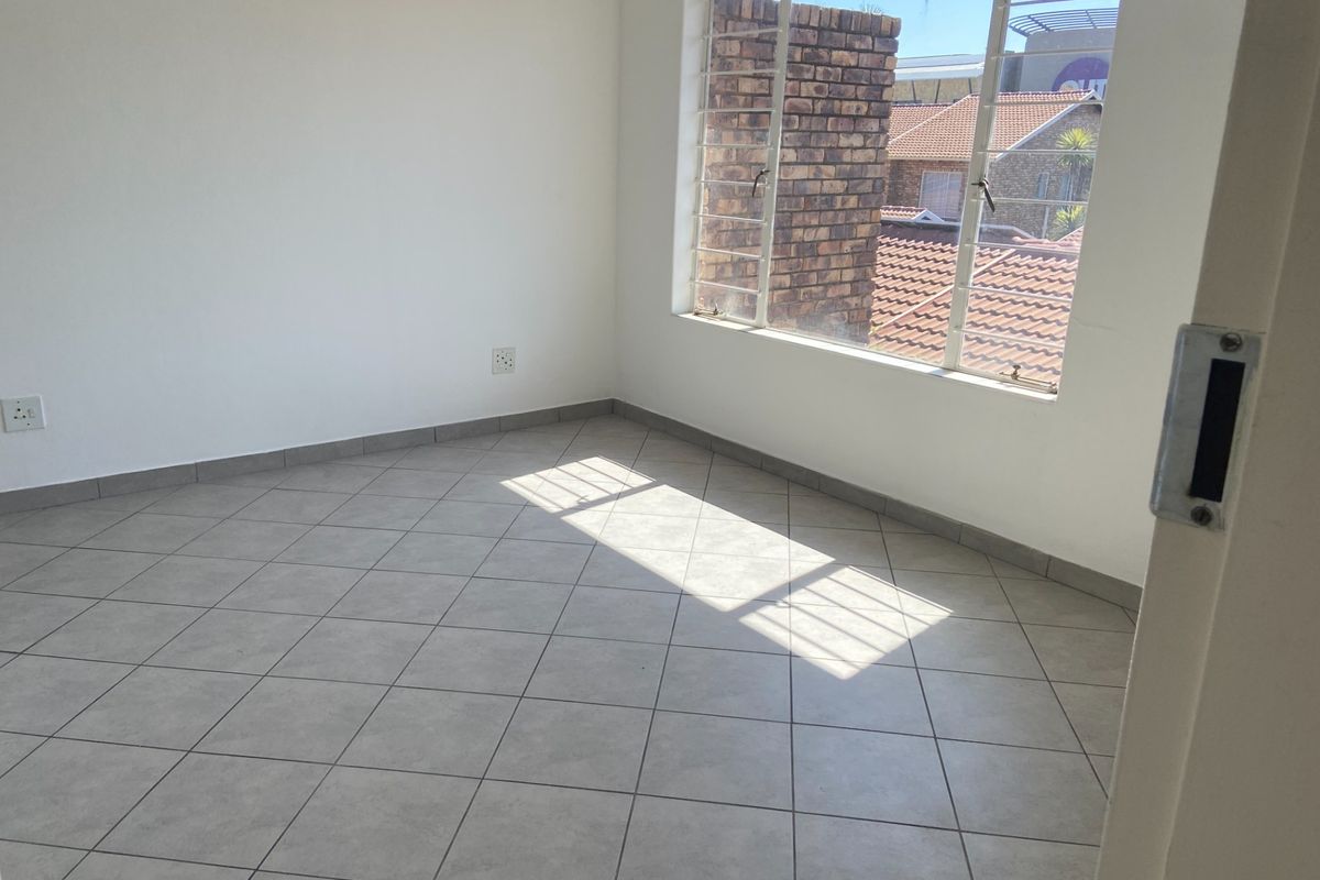 A bright, empty bedroom featuring neutral floor tiles, white walls, and a large window with security bars overlooking the neighborhood.