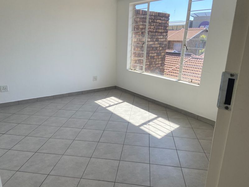 A bright, empty bedroom featuring neutral floor tiles, white walls, and a large window with security bars overlooking the neighborhood.