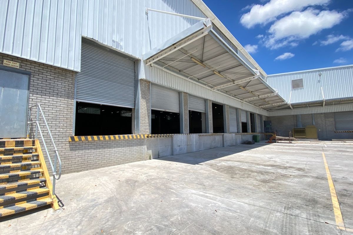 Industrial warehouse exterior featuring multiple roller shutter loading bays, a concrete loading dock, and a protective canopy over the service area.