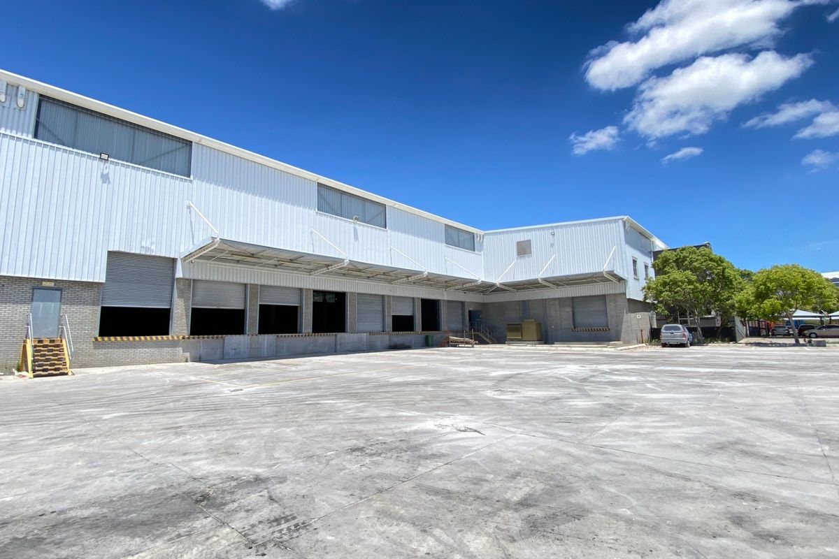 Industrial warehouse exterior featuring a large concrete loading yard, multiple roller shutter doors, and metal cladding with overhead canopy protection.