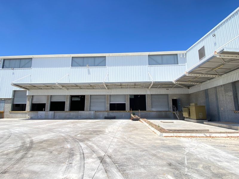 Industrial warehouse exterior featuring multiple loading docks with roller shutter doors, a large concrete yard, and protective metal canopy.