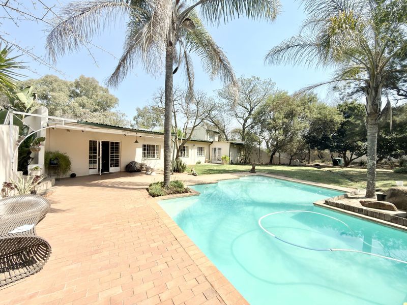 A spacious outdoor entertainment area featuring a swimming pool, brick-paved patio, and a covered veranda set within a mature garden.