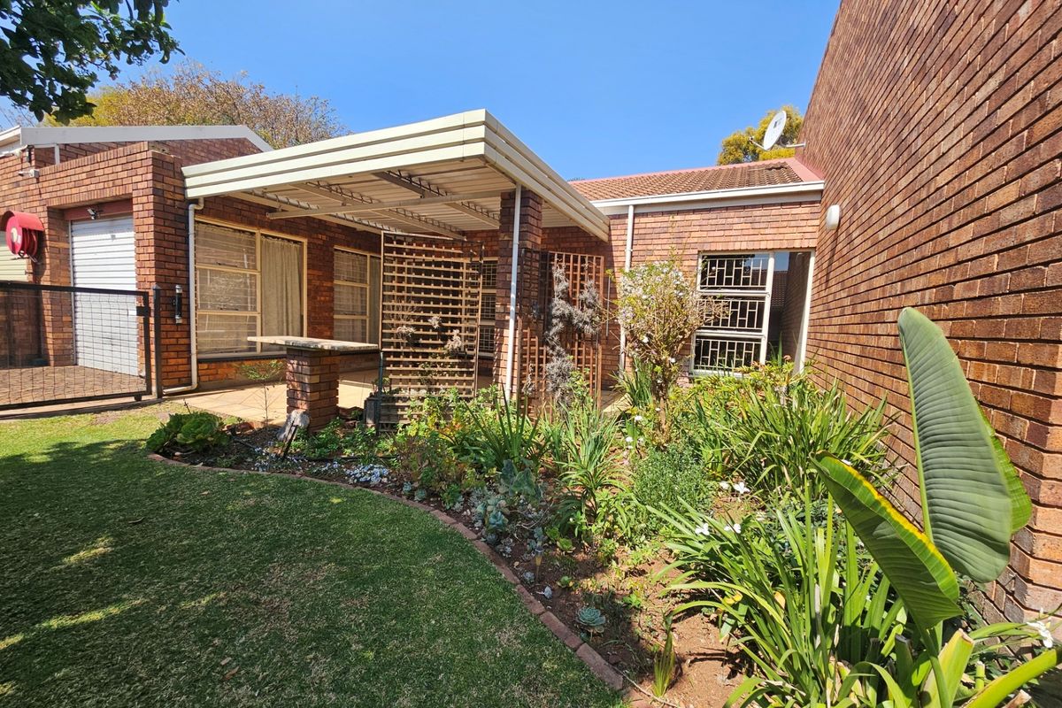 Rear exterior view of the townhouse featuring a brick facade, a covered patio area with a built-in braai, and a well-maintained garden.