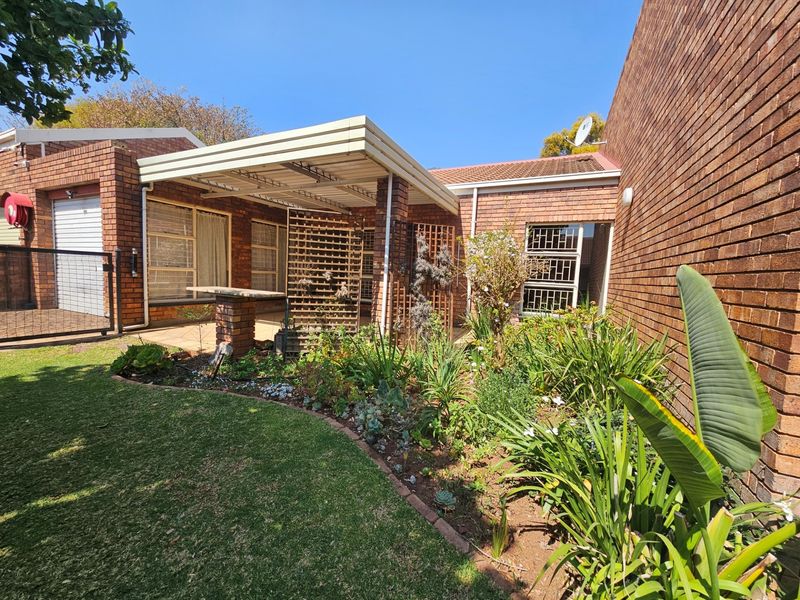 Rear exterior view of the townhouse featuring a brick facade, a covered patio area with a built-in braai, and a well-maintained garden.