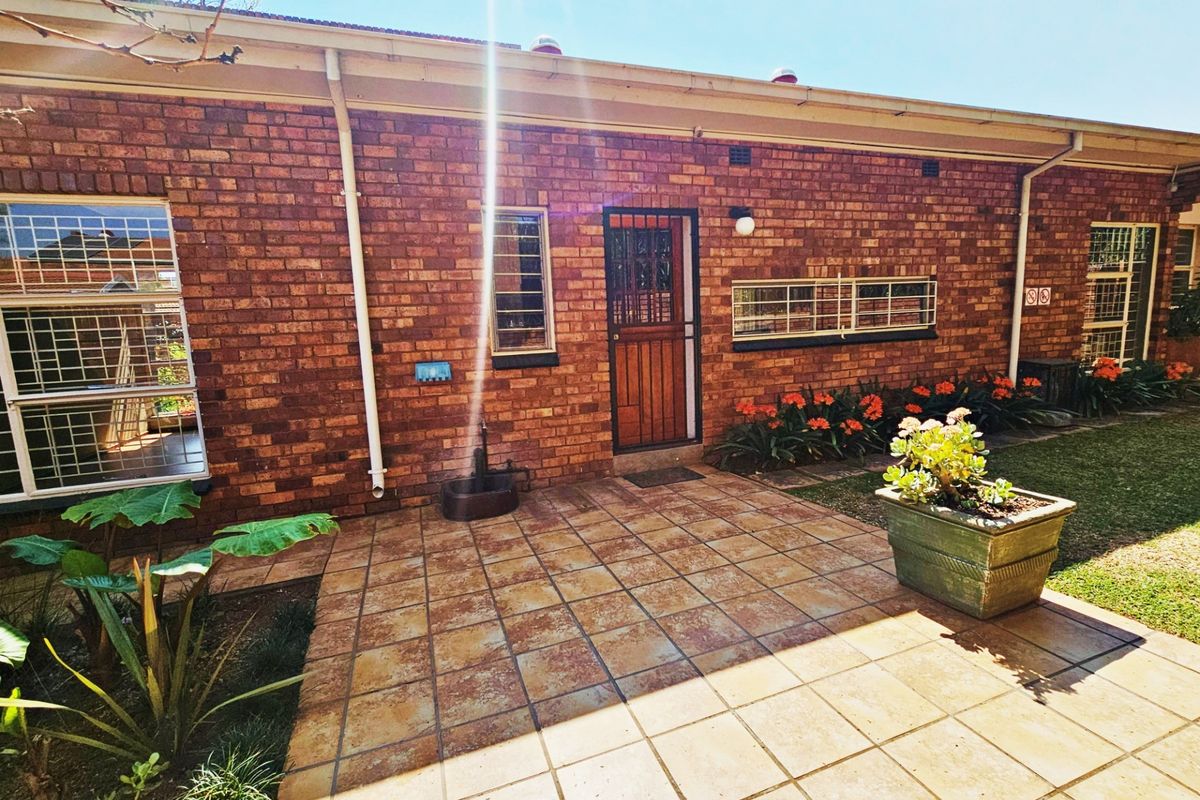 A rear exterior view of a brick townhouse featuring a tiled patio area, security bars on windows, and established garden beds.