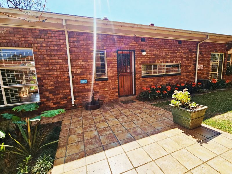 A rear exterior view of a brick townhouse featuring a tiled patio area, security bars on windows, and established garden beds.