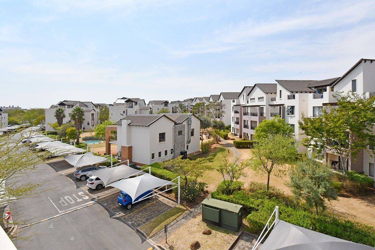 An elevated aerial view of a residential apartment complex featuring paved driveways, covered parking bays, and landscaped communal garden areas.