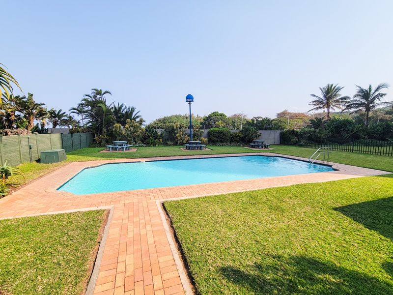 A well-maintained outdoor swimming pool area featuring a paved surround, manicured lawn, and secure perimeter fencing.