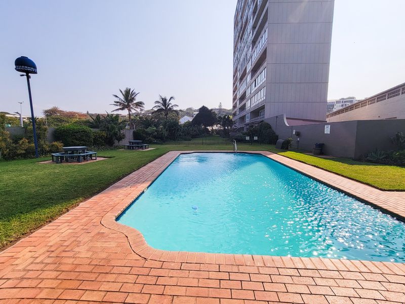 A well-maintained communal swimming pool area featuring brick paving, manicured lawn, and perimeter walling, set within a residential complex.