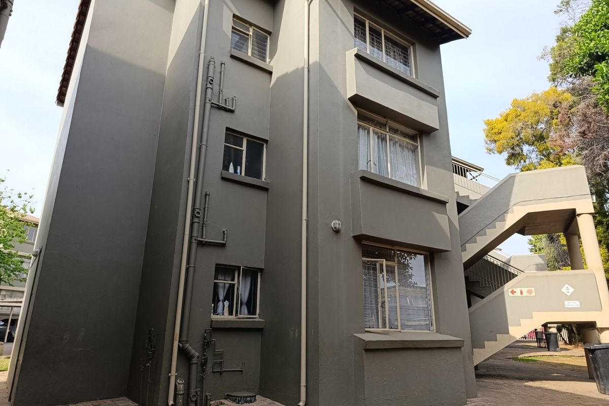 Exterior view of an apartment building featuring a paved walkway, external staircase, and exposed plumbing infrastructure.
