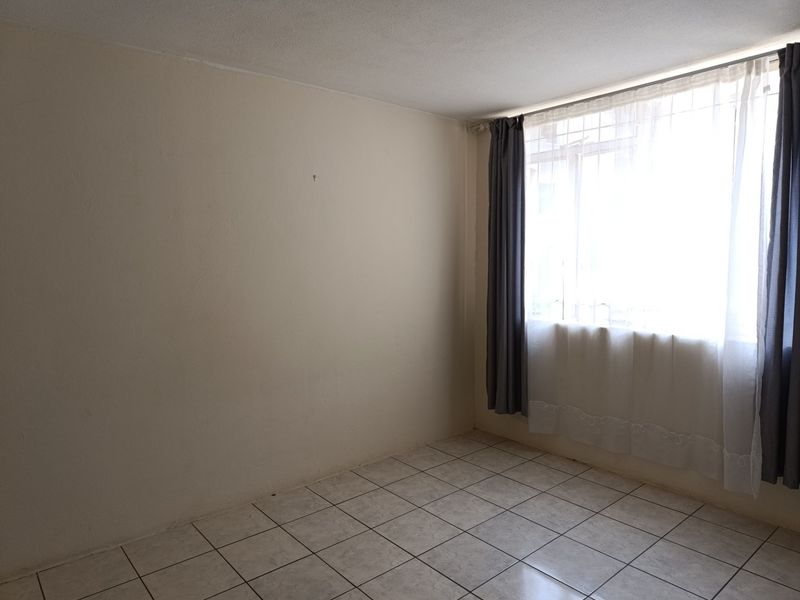 A bright, empty bedroom featuring neutral tiled flooring and a window fitted with security bars.