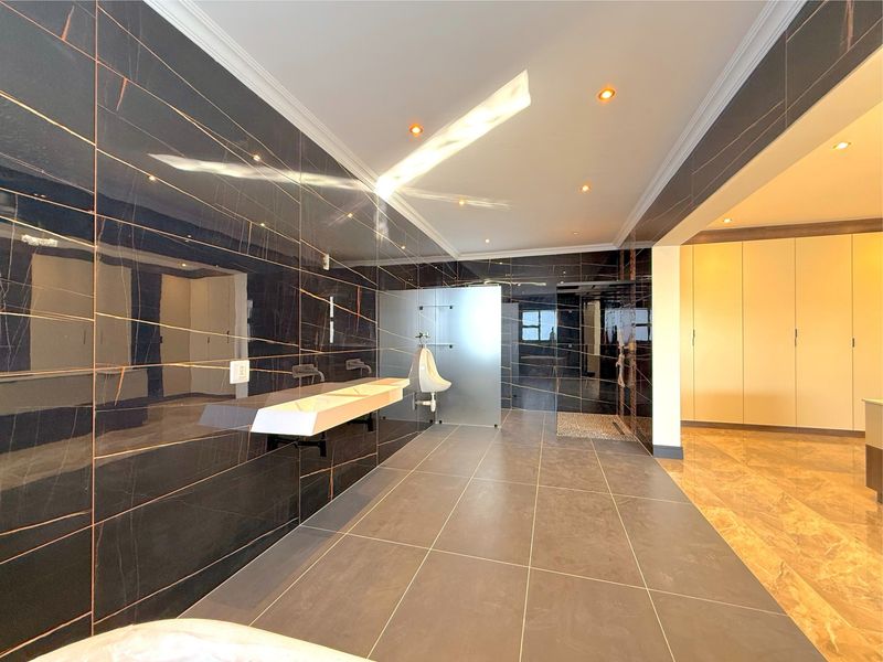 A modern, spacious bathroom featuring dark marble-effect wall tiles, a wall-mounted double vanity, a private urinal, and a walk-in shower area.