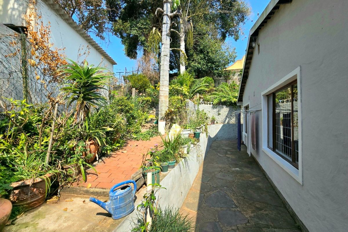 A rear garden area featuring a stone paved walkway alongside the house, a brick path, and a retaining wall with established landscaping.