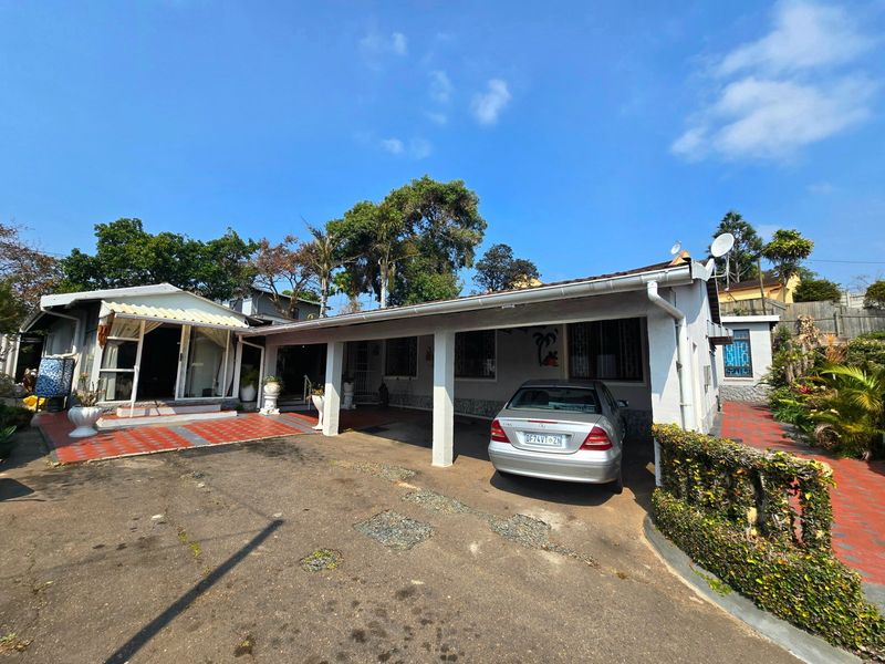 Front exterior view of the house featuring a covered carport, paved driveway, and tiled patio area with established garden surroundings.
