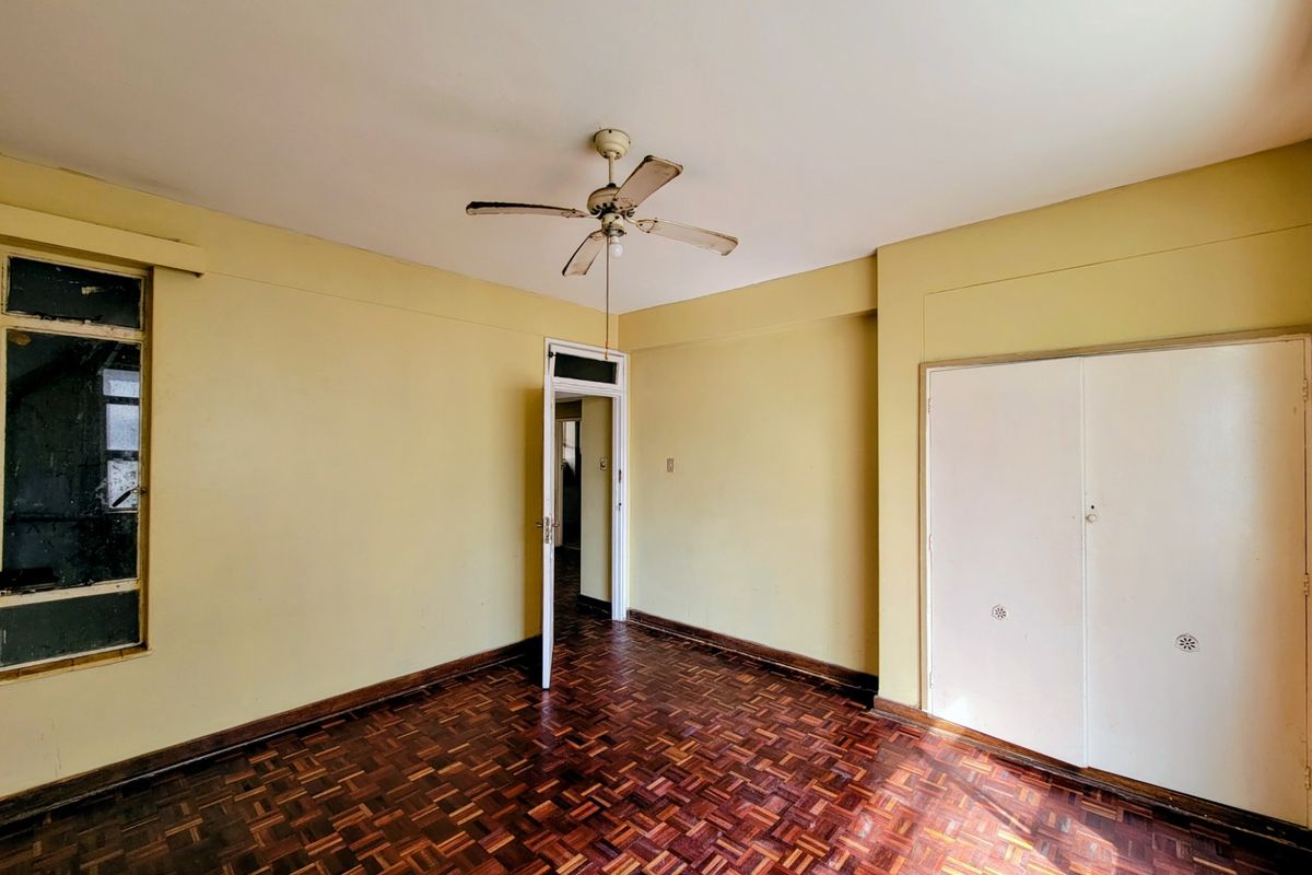 An empty bedroom featuring original parquet flooring, a ceiling fan, and built-in storage cupboards, requiring cosmetic updates to modernize the space.