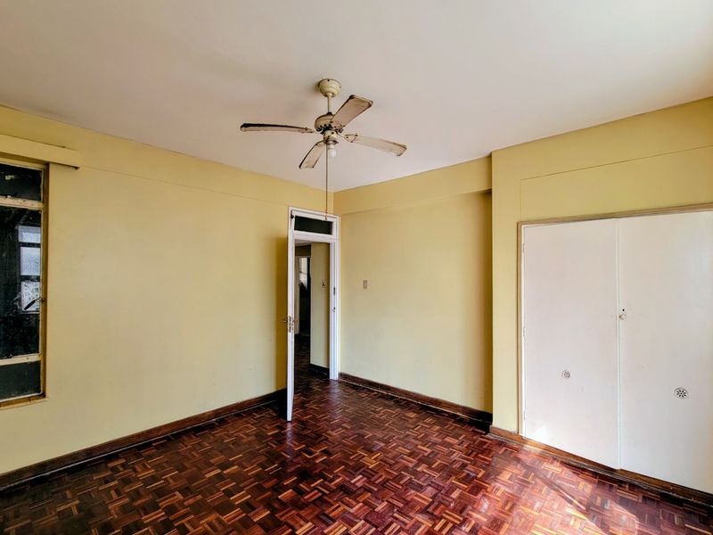 An empty bedroom featuring original parquet flooring, a ceiling fan, and built-in storage cupboards, requiring cosmetic updates to modernize the space.