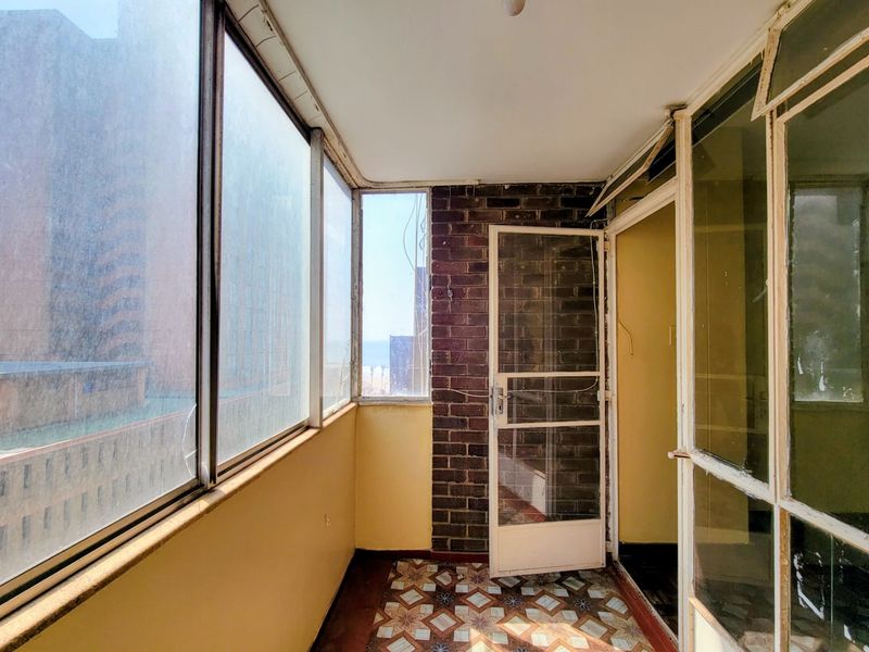 An enclosed balcony featuring a brick accent wall, tiled flooring, and large windows offering urban views, requiring some maintenance and cosmetic updates.