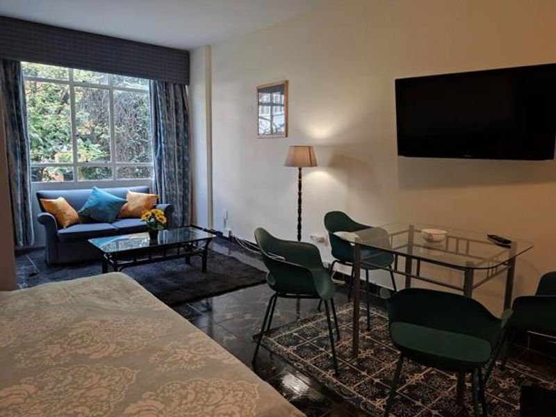 A living area featuring large windows, tiled flooring, and a wall-mounted television, providing a functional space for relaxation and dining.