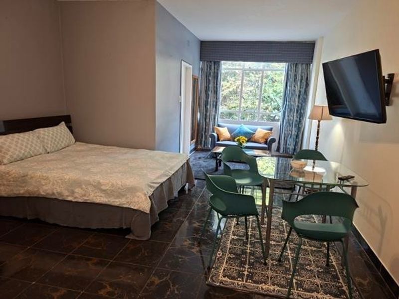 A spacious bedroom featuring dark tiled flooring, a large window with natural light, and a wall-mounted television.