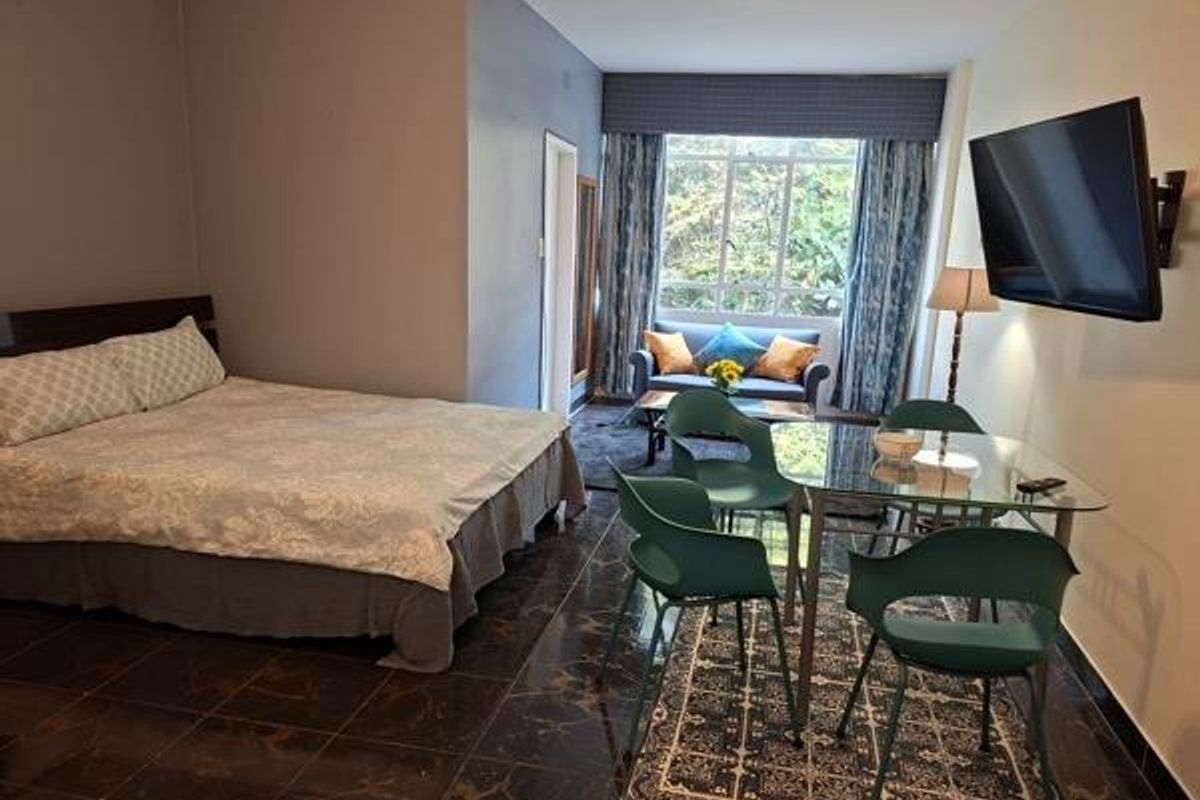 A spacious bedroom featuring dark tiled flooring, a large window with natural light, and a wall-mounted television.