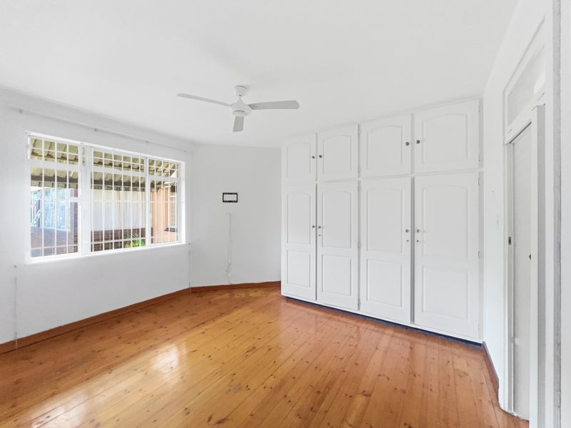 A bright, empty bedroom featuring polished timber flooring, ample built-in cupboard storage, and a ceiling fan.