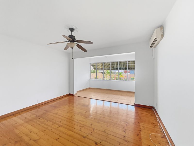 Spacious living area featuring polished timber flooring, a ceiling fan, split system air conditioning, and an adjoining tiled sunroom with security window bars.