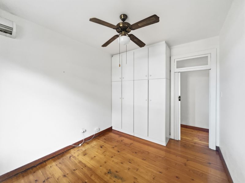 A bright, empty bedroom featuring polished timber flooring, a built-in wardrobe, a ceiling fan, and a wall-mounted air conditioning unit.
