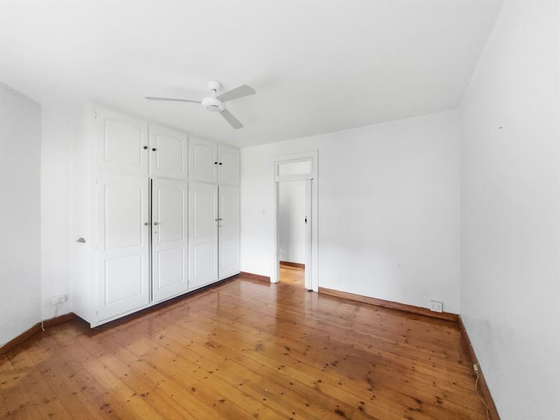 A bright, empty bedroom featuring polished timber flooring, a white built-in wardrobe, and a ceiling fan.