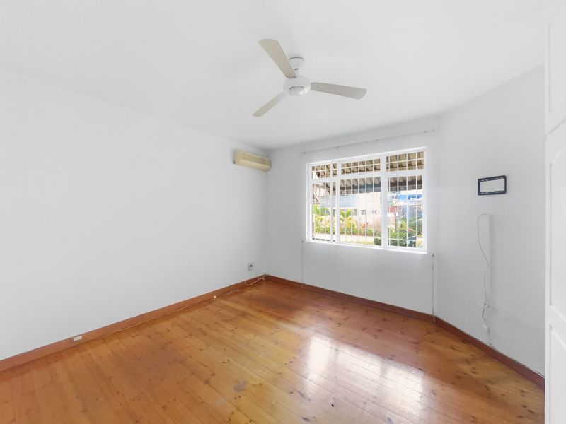 An empty bedroom featuring polished timber flooring, a ceiling fan, and a wall-mounted air conditioning unit with security bars on the window.