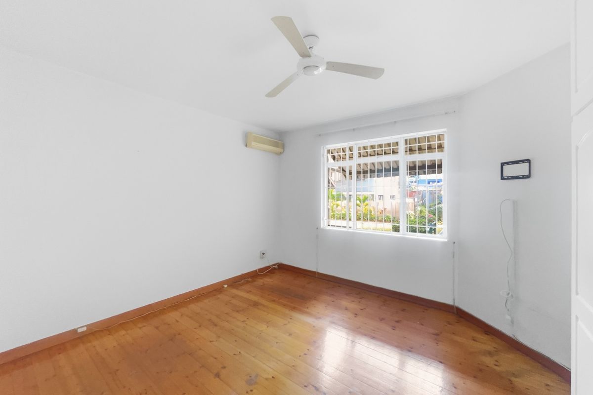An empty bedroom featuring polished timber flooring, a ceiling fan, and a wall-mounted air conditioning unit with security bars on the window.