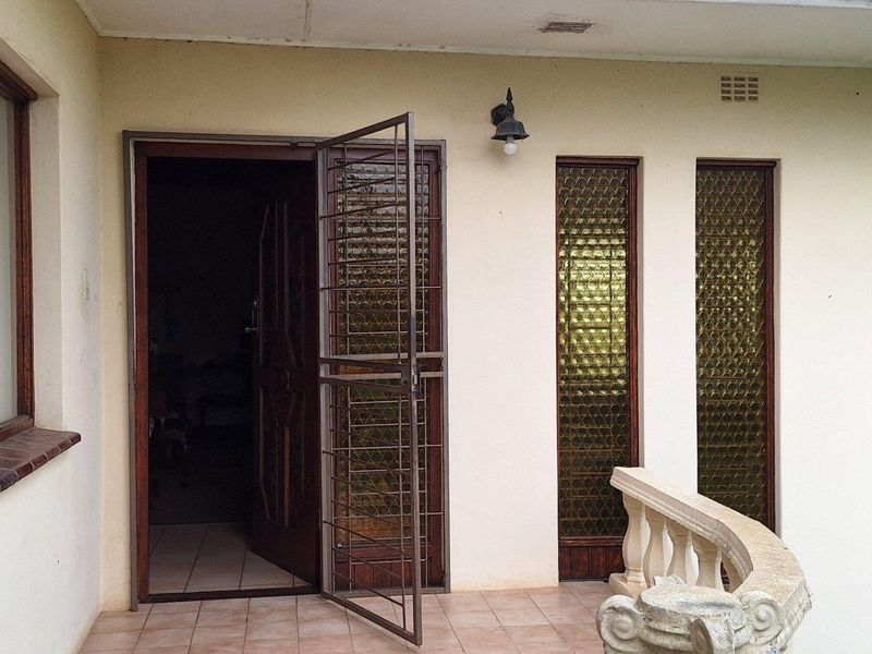 A covered patio area featuring tiled flooring, a decorative balustrade, and secure entrance access with timber-framed windows and textured glass detailing.