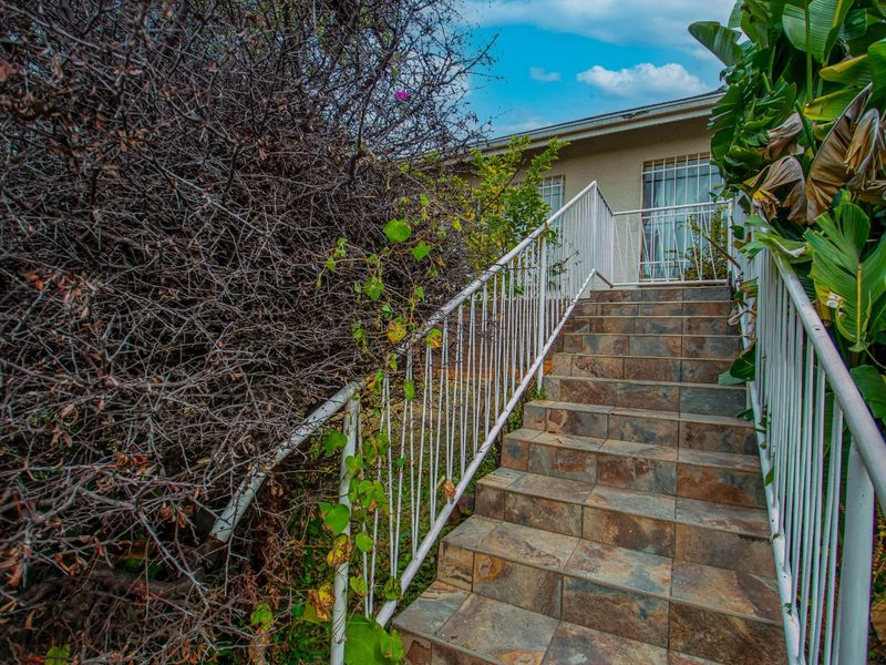 Exterior staircase with tiled steps and metal security railings leading to the property entrance, surrounded by overgrown garden foliage.