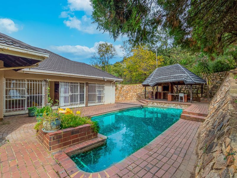 Outdoor pool area featuring a swimming pool, brick paved surround, and a covered gazebo with a built-in braai, set against a stone retaining wall.