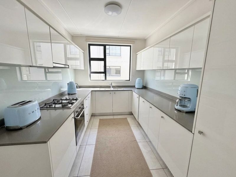 Modern galley-style kitchen featuring sleek white cabinetry, stone countertops, a gas hob, built-in oven, and a glass splashback.