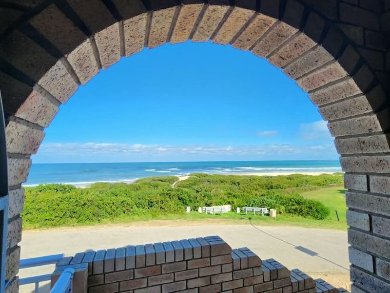 A scenic ocean view framed by a rustic brick archway and balustrade, overlooking coastal vegetation and a sandy beach.